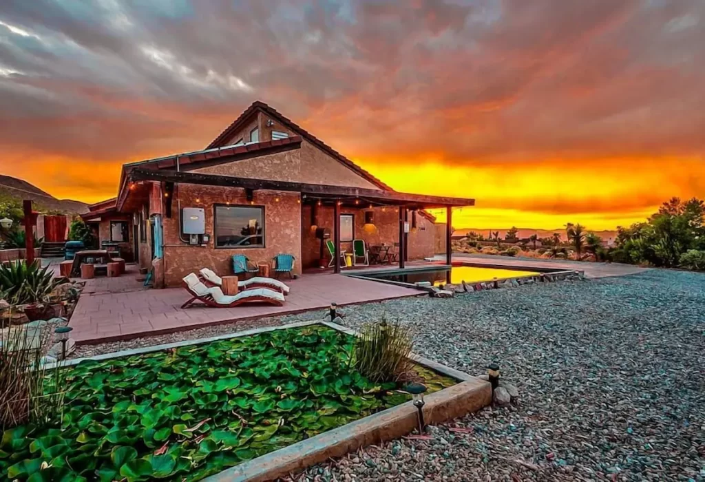 A beautiful desert home near Joshua Tree at sunset, featuring a spacious patio, swimming pool, and lush greenery in the foreground.