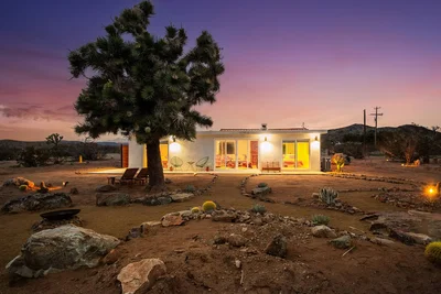 A desert home with large windows glowing at sunset, surrounded by rocky terrain and a prominent Joshua tree in the foreground.