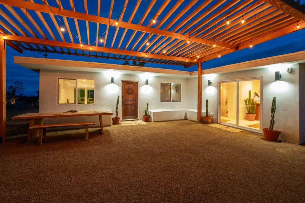 A cozy outdoor area of a modern desert home in Joshua Tree, featuring a wooden pergola with string lights, a large wooden bench, and potted cacti, set against a twilight sky.