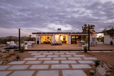 Exterior view of a modern desert home at sunset, featuring a spacious deck, outdoor seating, and decorative string lights.