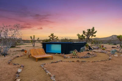 A modern desert home with a sleek black exterior and an inviting glass pool, set against a vibrant sunset sky in Joshua Tree, California. The scene features a wooden lounge chair, desert landscaping, and iconic Joshua trees in the background.