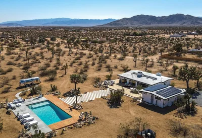 Aerial view of a desert property in Joshua Tree, featuring a swimming pool, hot tub, and a modern home surrounded by Joshua trees and mountain views.