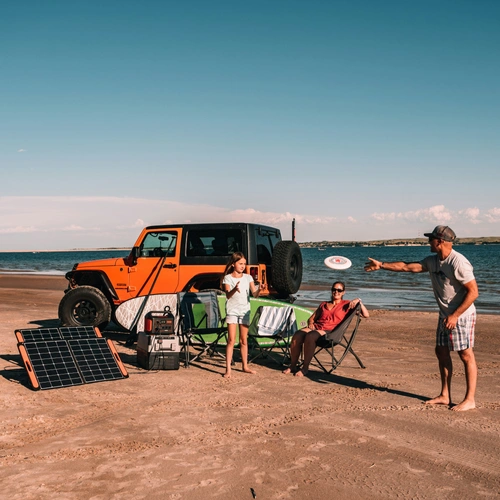 A family playing on a sandy beach next to an orange Jeep, using the Jackery Explorer 1000 v2 portable power station and solar panels to run a camping fridge.