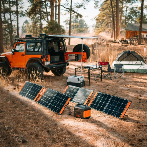 An overlanding campsite in a pine forest featuring an orange Jeep, with the Jackery Explorer 1000 v2 portable power station connected to four unfolded solar panels on the ground.