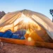 REI Anniversary Sale 2026 with Best Deals on Outdoor Gear. A camping tent pitched in a desert landscape during a golden hour sunrise, featuring a blue sleeping bag inside and a Joshua tree in the background.